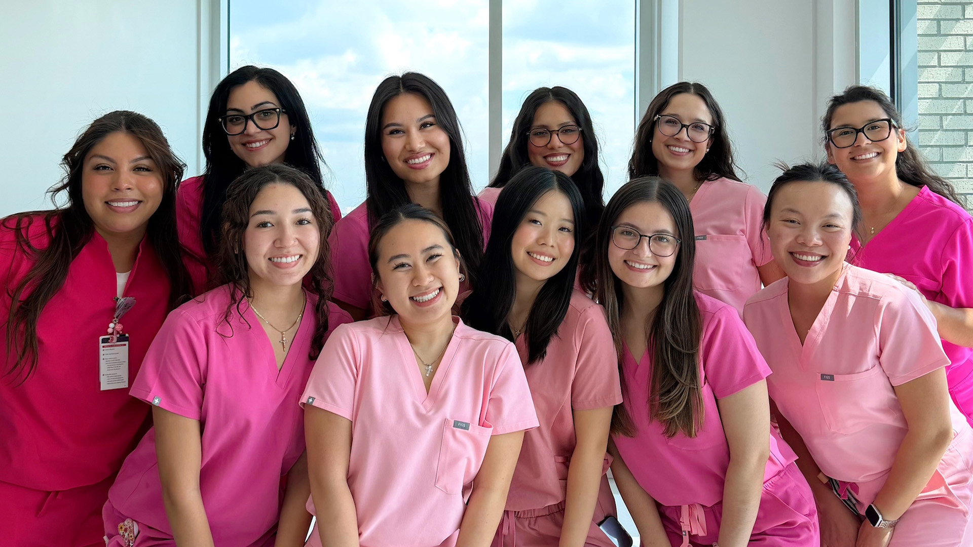 A group of people wearing pink scrubs pose together in front of large windows.
