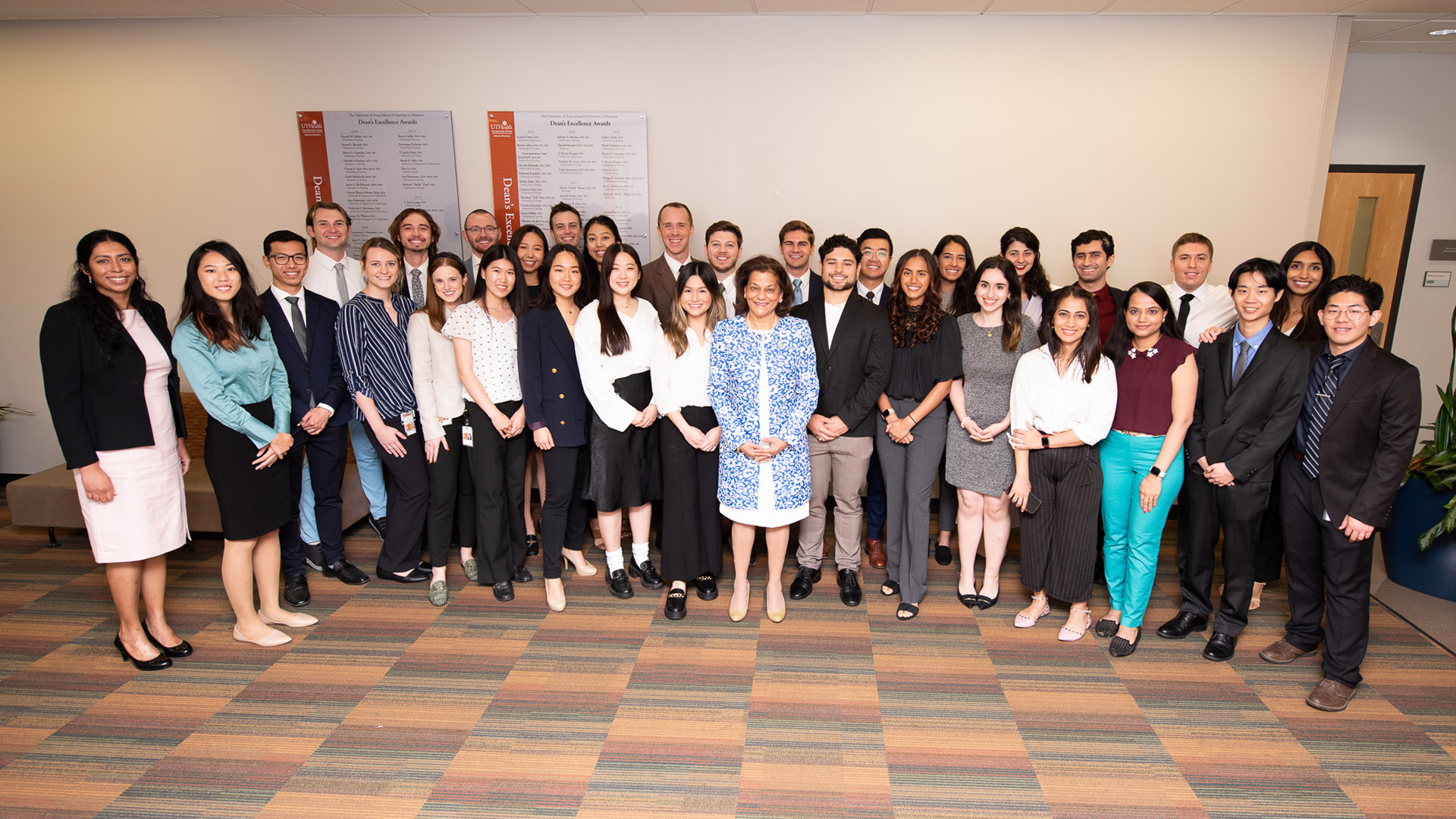 A large group of about people stand together indoors in business or business?casual attire, smiling toward the camera in a professional setting with neutral walls, patterned carpet, and plants.