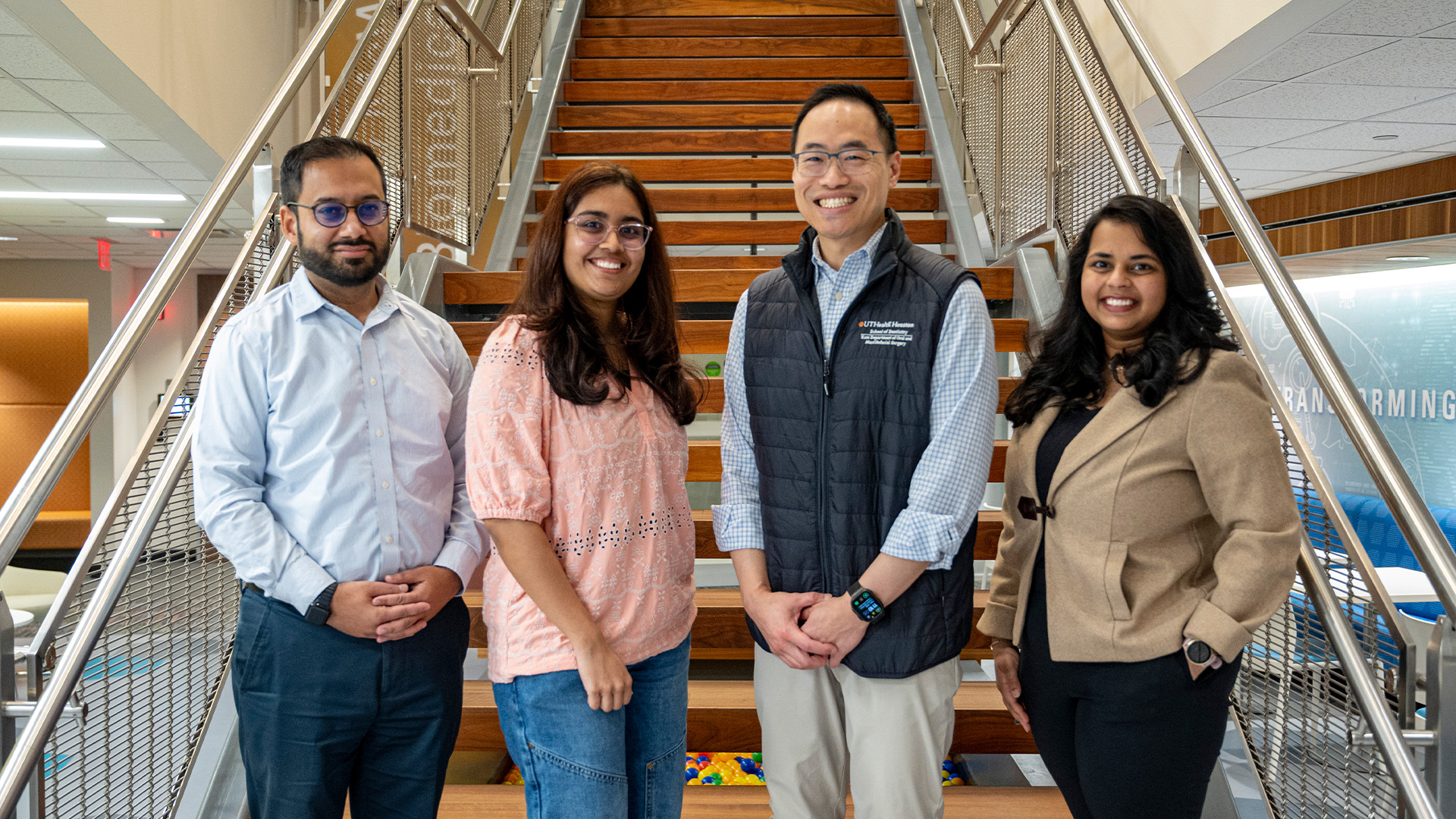 Four people, dressed in business?casual attire, stand on a wide wooden staircase inside a modern building, smiling toward the camera.