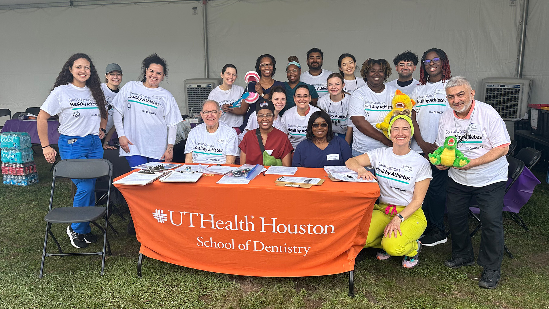 A group of people pose under a large white tent behind a table with an orange UTHealth Houston School of Dentistry tablecloth. Most wear “Special Olympics Healthy Athletes.”