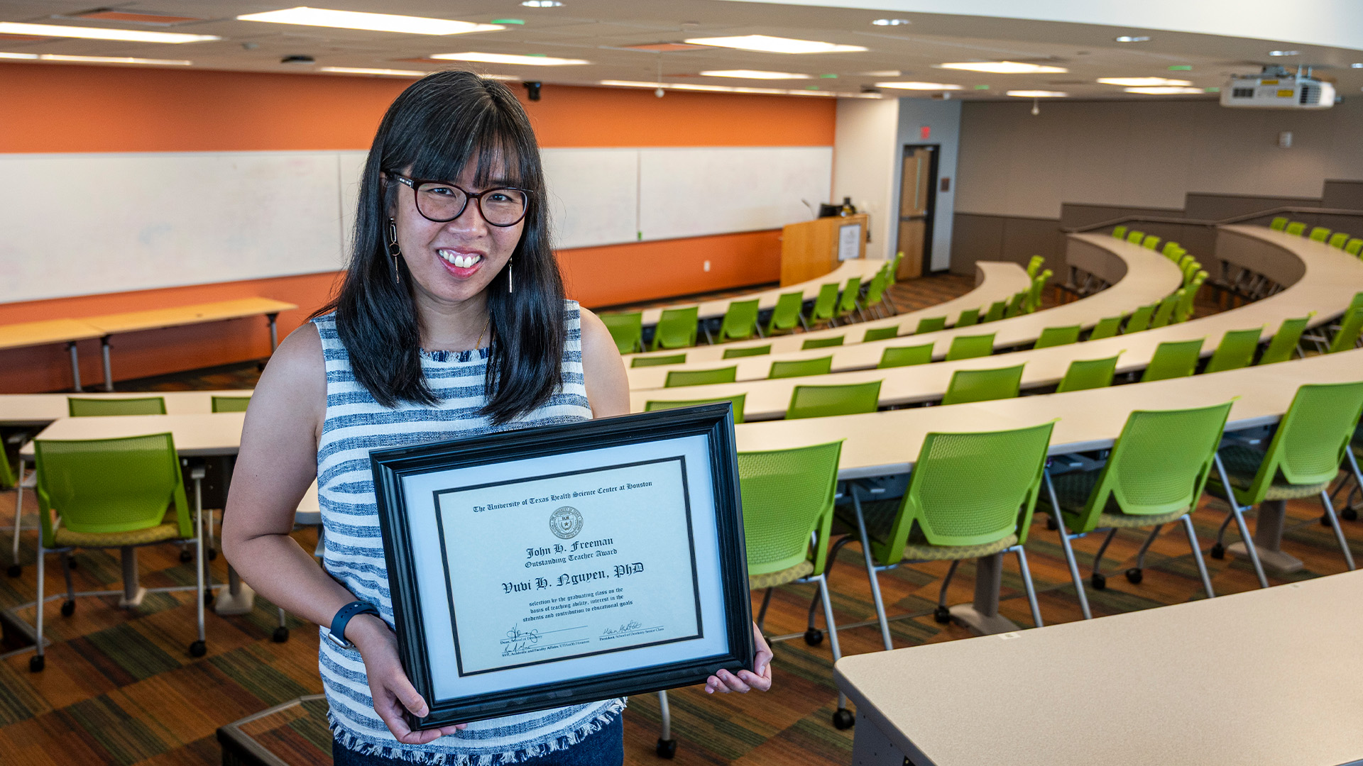 Person holding a framed certificate while standing in a classroom with tiered green seating and a projection screen in the background.