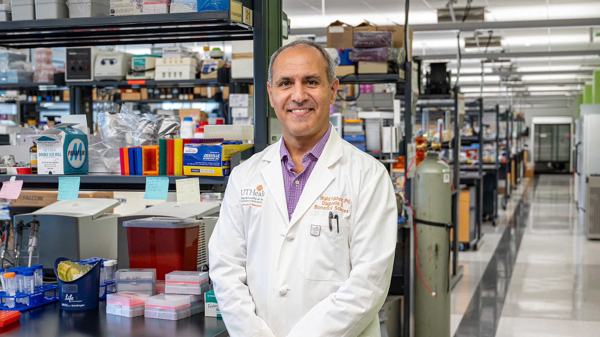 A person in a white lab coat stands in a well-equipped research laboratory with shelves of supplies, pipette tip boxes, test tube racks, and other scientific equipment in the background.