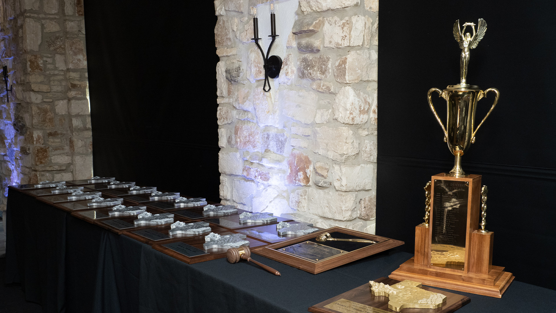 A black?covered table holds several awards against a stone wall, with rectangular wooden Texas?themed plaques on the left and a large gold trophy with a winged figure on a wooden base on the right.